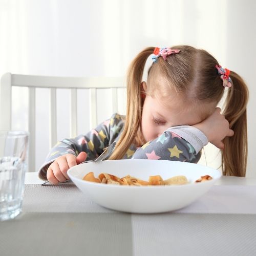 A small girl asleep at the breakfast table, a bowel of cereal is in front of her