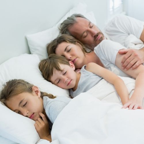 A father and mother are sleeping in a bed with their 2 children. All of the linens are white.ng in a bed