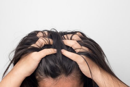 A girl with brown hair has both of her hands in her hair touching her scalp pulling her hair out.